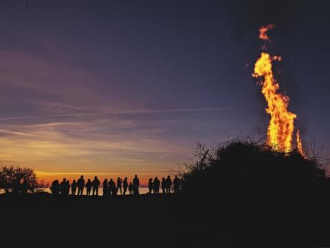 Menschen stehen um ein Lagerfeuer bei Sonnenuntergang, während die Sonne am Horizont untergeht und den Himmel färbt.